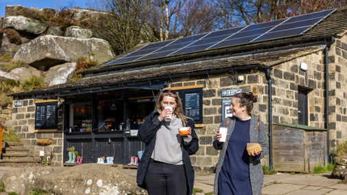 Two women walk away from a stone-built cafe enjoying a warm drink as they go with blue autumnal skies above